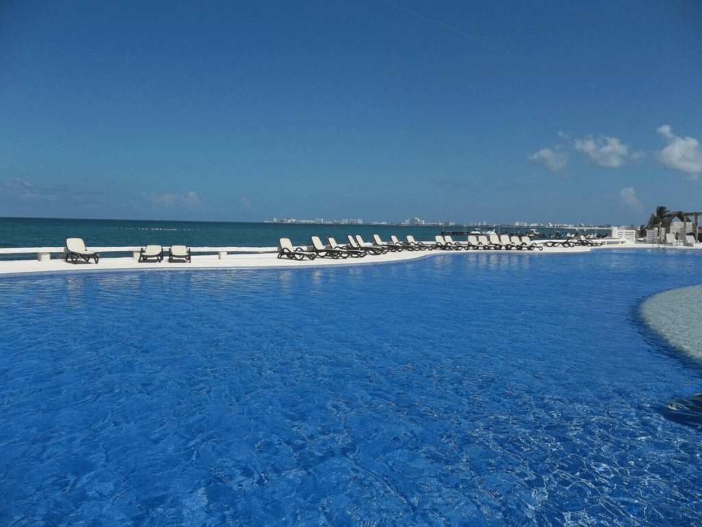 A beautiful pool area with clear blue water, lined with lounge chairs, overlooking the ocean under a bright blue sky.