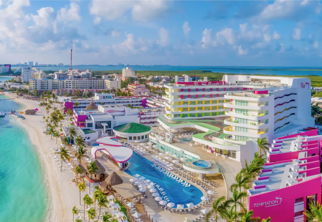 Aerial view of a vibrant resort complex near a turquoise beach, featuring modern architecture, palm trees, and a swimming pool.