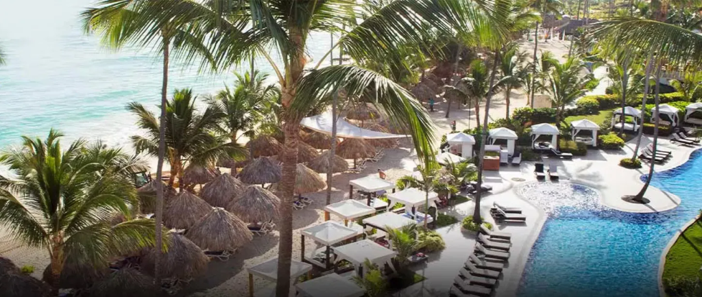 Aerial view of a tropical beach resort featuring palm trees, thatched cabanas, and a swimming pool next to the ocean.